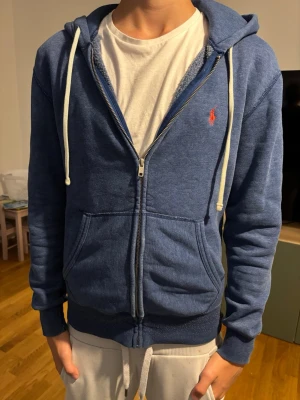 Blå zip hoodie med vit dragsko - Snygg blå hoodie med dragkedja och vit dragsko. Klassisk design med två stora fickor framtill och huva. Liten röd logga broderad på bröstet. Perfekt till chill vardagsstil och passar till det mesta. Mjuk och skön bomullsblandning.