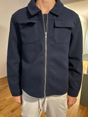 Mörkblå overshirt med dragkedja - Stilren mörkblå overshirt med rak passform och två bröstfickor med lock. Jackan har en klassisk krage och stängs med silverfärgad dragkedja framtill. Materialet ser ut att vara ullblandning, vilket ger en mjuk och varm känsla. Perfekt lager till hösten.