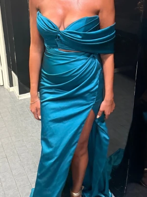 Turkosblå klänning från kosovo - Säljer en elegant turkos långklänning med offshoulder och omlott-detalj i midjan. Klänningen har en hög slits framtill och är tillverkad i ett glansigt satin material som ger en lyxig känsla. Perfekt för speciella tillfällen. Den är köpt från Kosovo. pris kan diskuteras. Använt endast en kväll!