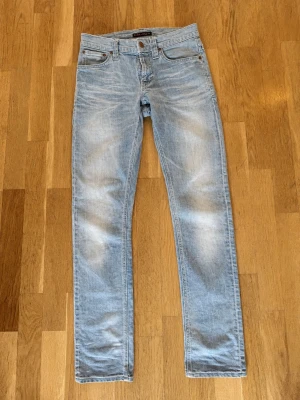 Nudie Jeans  - Säljer ett par ljusblå jeans från Nudie med raka ben och klassisk femficksmodell. Jeansen har slitningar och en avslappnad tvättad look. Tillverkade i bomull med metallknappar och nitar. Perfekta för en casual stil.