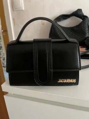 Svart Jacquemus Le Chiquito väska - Säljer en svart Jacquemus Le Chiquito handväska i slätt läder med guldfärgad logga framtill. Väskan har ett topphandtag, lock med magnetstängning och en stilren, boxig form. Perfekt för dig som gillar trendiga accessoarer och minimalistisk design. Den är köpt på Spaniens marknad!! Bandet följer även med