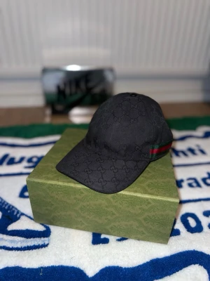 Svart Gucci keps med monogram - Snygg svart keps från Gucci med klassiskt GG-monogram över hela kepsen och en grön-röd rand på sidan. Kepsen har böjd skärm och justerbart band bak. Tillverkad i canvasmaterial som ger en exklusiv känsla.