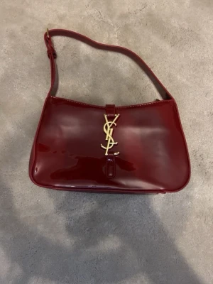 Vinröd handväska YSL - Snygg vinröd handväska i lackat material från YSL med guldigt logospänne framtill. Väskan har en justerbar axelrem och stilren, rektangulär form. Perfekt accessoar för att lyfta din outfit.