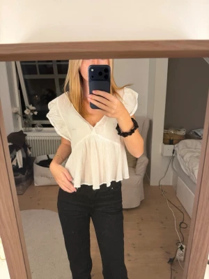 Vit blus med volang och v-ringning - Söt vit blus med v-ringning, volangdetaljer och korta ärmar. Blusen har en luftig passform och knytning i ryggen. Perfekt till jeans eller kjol för en trendig look.
