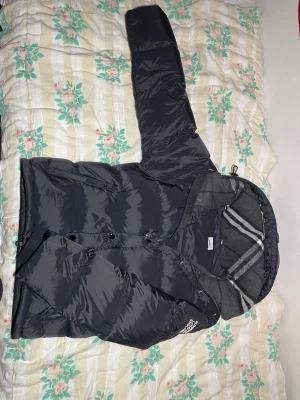 Burberry puffer  - Snygg svart pufferjacka från Burberry med stor logga på ärmen och klassiskt rutigt foder i huvan. Jackan har quiltad design, dragkedja och knappar framtill samt en mysig huva. Perfekt för kalla dagar och riktigt trendig look. Perfekt för kalla vintern som är påväg, kom gärna med bud eller frågor om ni har några som helst funderingar. Jackan har några riktigt schyssta detaljer som ej många jackor har🙌