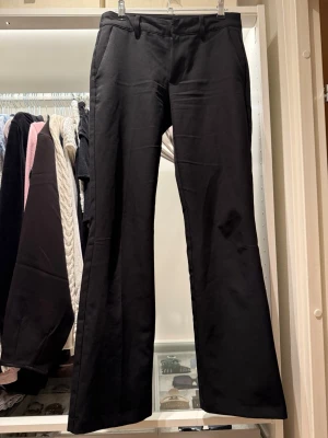 Svarta utsvängda kostymbyxor - Stilrena svarta kostymbyxor med utsvängda ben och klassiska fickor fram. Byxorna har hällor för skärp och en enkel design som passar till många olika looks. Materialet är en mjuk och följsam textil med lätt stretch. Inga skavanker alls. Inrebenslängd 74,5cm midjemått 34,5cm skev till midja 21,5cm 
