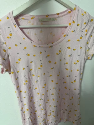 Ljusrosa t-shirt med citronmönster - Söt ljusrosa t-shirt med små gula citroner över hela tyget. T-shirten har rund halsringning och korta ärmar. Perfekt för dig som gillar fräscha och lekfulla plagg med somrig känsla. Använd få gånger!!