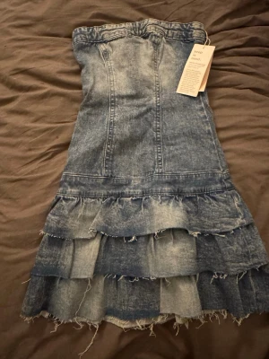 Blå jeansklänning med volanger - Säljer en blå jeansklänning med flera lager volanger nedtill. Klänningen är axelbandslös och har en fin, rå kant längst ner. Perfekt för dig som gillar denim och vill sticka ut med en unik look. Helt ny oanvänd med prislapp