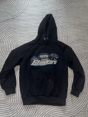 Svart hoodie Trapstar London Shooters - Svart hoodie med stor huva och trycket 'Trapstar London Shooters' i vitt, blått och grått framtill. Hoodien har känguruficka och ribbade muddar vid ärmar och nederkant. Perfekt för en streetwear-look