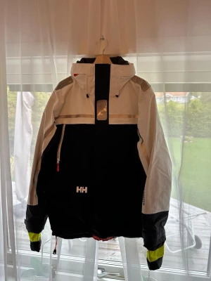 Helly Hansen svartvit vindjacka - Segeljacka från Helly Hansen i svart och vitt med reflekterande detaljer och dragkedja på bröstet. Jackan har luva som man kan stoppa in, hög krage och logga både fram och bak. Ärmsluten har svarta och neongula partier för extra synlighet som är innanför jackan som man kan dra ut om man vill. Fickor med fleecefoder.