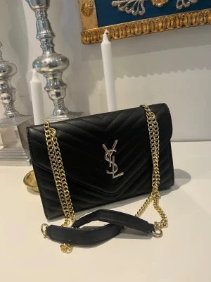 Svart axelväska från Saint Laurent - Snygg svart axelväska replika från Saint Laurent med guldfärgad kedjerem och ikonisk YSL-logga framtill. Knappast använd och är fortfarande i fint skick. 25x17 cm