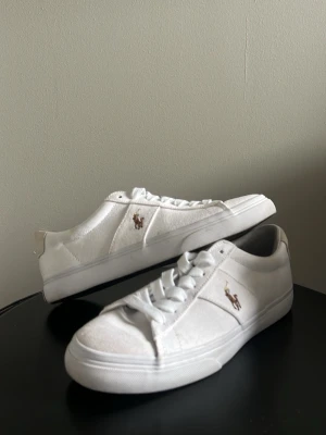 Vita låga sneakers från Polo Ralph Lauren - Stilrena vita sneakers från Polo Ralph Lauren med klassisk låg profil och broderad logga på sidan. Skorna har ovandel i canvas och detaljer i läder, samt vita snören och sula. Perfekta för en clean och fräsch look. Storlek 41