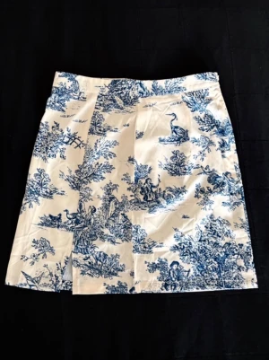 Vit och blå kortkjol med minimalistisk slit - Vit kortkjol från Cider med blått, detaljrikt mönster i vintage-stil. Kjolen har en rak passform och stängs med dragkedja i sidan. Tillverkad i ett mjukt material som känns bekvämt mot huden.