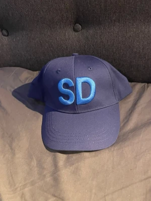 Mörkblå keps med SD brodyr - Snygg mörkblå keps med stora broderade bokstäver 'SD' framtill i blått. På sidan finns en liten broderad bi tillsammans med texten 'SD'. Justerbar baktill för perfekt passform. Klassisk formad skärm och ventilerande hål upptill.