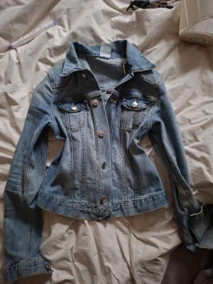 Ljusblå croppad jeansjacka - Trendig ljusblå croppad jeansjacka med markerade sömmar och klassiska bröstfickor med lock. Jackan har silverfärgade knappar framtill och på fickorna samt en figurnära passform som framhäver midjan. Perfekt för en cool och avslappnad stil.