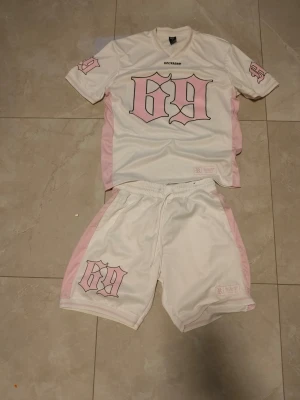 Vitt och rosa set med tryck 69 - Säljer ett tvådelat set med t-shirt och shorts i vitt med ljusrosa detaljer. Både tröjan och shortsen har stort tryck med siffran 69 i rosa och svart. Setet har sportig look med v-ringad hals och elastisk midja på shortsen. Perfekt för dig som gillar streetwear.