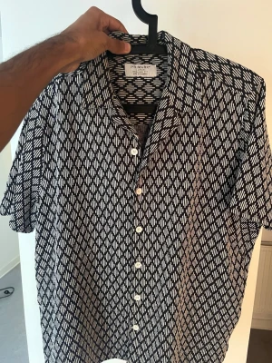Mönstrad kortärmad skjorta från Primark | Strl L - Säljer en riktigt snygg kortärmad skjorta från Primark i storlek L. Skjortan har ett unikt svartvit geometrisk/geometriskt mönster och en skön resort-krage. Perfekt att slänga på sig över ett linne eller bär som den är under varma dagar!  Märke: Primark  Storlek: L (Large)  Passform: Regular Fit  Skick: Mycket gott skick! 🛍️ Samfrakta & spara pengar! Kika gärna på mina andra annonser på profilen! Säljer flera andra snygga plagg just nu. ✨ Paketrabatt: Jag erbjuder självklart paketrabatt om du vil