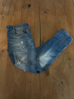 Blå slitna jeans med detaljer - Säljer ett par blå jeans med slitningar och distressed detaljer på både fram- och baksida. Jeansen har klassisk femficksmodell, normal midja och raka ben. Materialet är denim i bomull och de har en snygg tvättad look med ljusare partier.