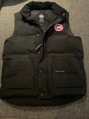 Svart dunväst från Canada Goose - Snygg svart dunväst från Canada Goose med hög krage, två stora framfickor och klassisk logopatch på bröstet. Västen har dragkedja och tryckknappar framtill samt diskret Canada Goose-märke på fickan. Perfekt för lager-på-lager och kyliga dagar.
