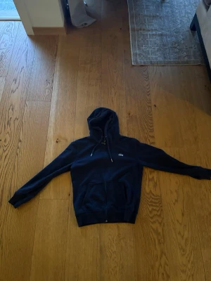 Mörkblå hoodie med dragkedja - Snygg mörkblå hoodie med dragkedja och huva. Modellen har fickor framtill och justerbara snören i huvan. Perfekt för en avslappnad och stilren look. Liten logga på bröstet ger en clean touch.