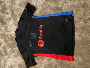 Svart FC Barcelona matchtröja - Säljer en svart FC Barcelona matchtröja med röda och blå detaljer på sidorna. Tröjan har tryck med Spotify-logga framtill, Nike-logga och klubbmärke. På baksidan finns UNHCR-tryck. Perfekt för dig som älskar fotboll och vill ha en snygg supportertröja.