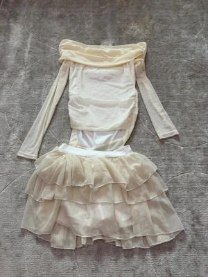 Off shoulder cremefärgad volangklänning - Trendig cremefärgad klänning med off shoulder-design och långa ärmar. Kjolen har flera lager volanger i tunt, skirt tyg som ger en romantisk känsla. Överdelen är figurnära och lätt rynkad för en snygg siluett. Perfekt för dig som gillar en feminin och unik stil.