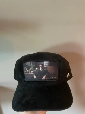 Svart keps från Maggiore med motiv - Snygg svart keps från Maggiore med mesh på sidorna och ett unikt motiv framtill som föreställer en man i kostym. Kepsen har böjd skärm och justerbar baksida. Perfekt för dig som vill sticka ut med en cool accessoar.