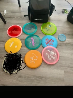 Disc golf set med väska - Säljer ett set med discgolfdiscar i olika färger och former samt en svart Latitude 64° discgolfväska. Discarna har tryck i olika färger och märken, bland annat gul, röd, blå, grön och rosa. Perfekt för dig som vill börja eller utveckla ditt discgolfspel. 8 discar och en väska medföljer