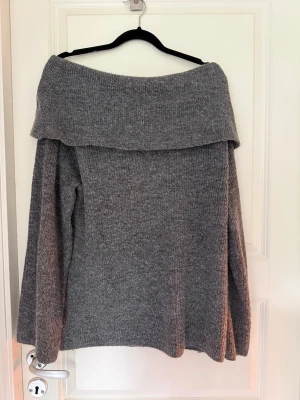 Grå off shoulder stickad tröja - Mysig grå stickad tröja med bred off shoulder-krage och långa ärmar. Tröjan har en avslappnad passform och är perfekt för kyliga dagar. Materialet känns mjukt och behagligt mot huden. Enkel att matcha med jeans eller kjol.