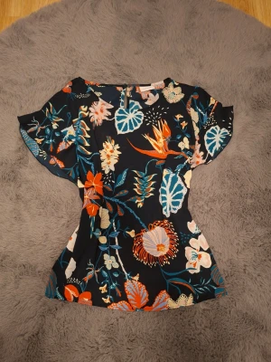 Blommig blus från Vila - Snygg blus från Vila med mörkblå bas och färgglatt blommönster i orange, blått, vitt och beige. Blusen har korta, vida ärmar och rund halsringning. Materialet känns lätt och luftigt, perfekt för en trendig look.