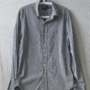 Randig skjorta från Polo Ralph Lauren - Snygg skjorta från Polo Ralph Lauren i svart och vitt randigt mönster. Klassisk krage, långa ärmar och knappar framtill. Custom fit-modell med diskret broderad logga i lila på bröstet. Perfekt för en stilren look.
