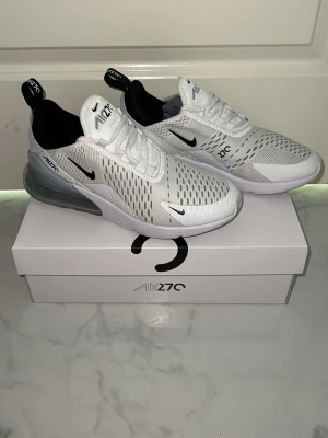 Nike Air Max 270 vita sneakers strl 40 - Nike Air Max 270 sneakers i vitt med svarta detaljer och synlig luftbubbla i hälen. Ovandelen är i mesh för bra ventilation och har en modern, sportig look. Swoosh-logga på sidan och dragflik bak. Perfekta för dig som gillar stilrena och bekväma sneakers.