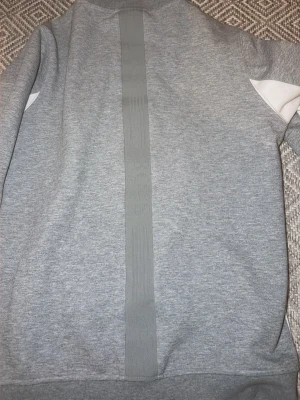 Grå ziptröja med vita detaljer - Snygg grå ziptröja med vita paneler på sidorna och mörkgrå detaljer. Tröjan har en rak passform, ribbade muddar och dragkedja framtill. Liten logotyp på bröstet och ståkrage för en sportig look. Perfekt till vardags eller träning.