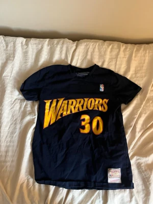 Warriors Curry 30 t-shirt i mörkblått - Snygg mörkblå t-shirt med stort gult Warriors-tryck och nummer 30 på framsidan. På baksidan står det Curry och 30 i samma färg. Perfekt för basketfans som vill visa sitt stöd för laget.
