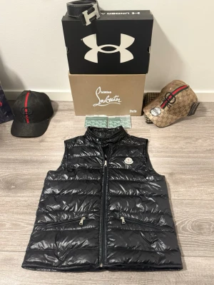 Moncler gui - INGA SKADOR, VÄLDIGT BRA SKICK.           Snygg svart dunväst från Moncler med glansig finish och quiltad design. Västen har två dragkedjeförsedda fickor framtill och en hög krage. Moncler-logga på bröstet ger en exklusiv touch. Perfekt för lager-på-lager och streetwear.