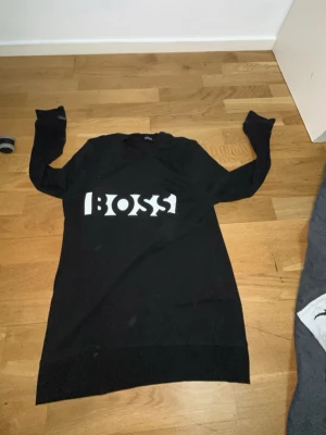 Svart långärmad tröja med BOSS-tryck - Snygg svart långärmad tröja med stort vitt BOSS-tryck på bröstet. Tröjan har en rak passform och är tillverkad i ett mjukt material som passar perfekt till en avslappnad stil. Enkel design med ribbade muddar vid ärmslut och nederkant.