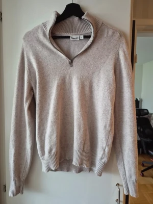 Beige halvzip tröja från Kappahl - Mysig beige stickad tröja från Kappahl med halvzip och hög krage. Tröjan har långa ärmar och ribbade muddar vid ärmslut och nederkant. Perfekt för lager på lager och enkel att matcha med jeans eller kjol.