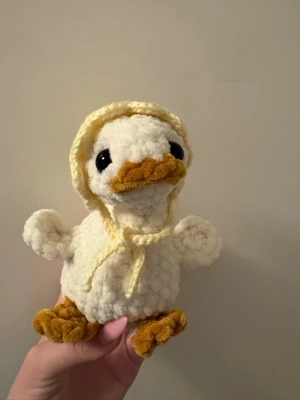 Virkad gås med huva - Supergullig handgjord virkad gås i mjukt vitt garn med gula detaljer på näbb, fötter och en söt huva. Gåsen har stora svarta ögon och fluffig struktur, perfekt som dekoration eller gosedjur.