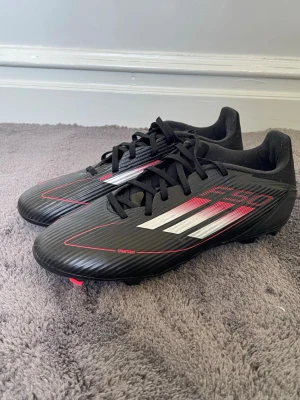 Svarta Adidas F50 fotbollsskor - Säljer ett par svarta Adidas F50 fotbollsskor med röda och vita detaljer. Skorna har klassisk snörning, låg profil och slitstark syntetisk ovandel. Perfekta för spel på både natur- och konstgräs. Snygg design med de ikoniska tre ränderna på sidan.