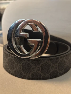Svart GG-bälte med silverdetalj - Snyggt svart bälte med klassiskt GG-mönster och stor silverfärgad spänne i metall. Bältet har en stilren design och är tillverkat i skinn med präglat mönster längs hela remmen. Perfekt accessoar för att lyfta din outfit.