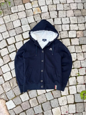 Mörkblå hoodie med knappar från Jean Paul - Mörkblå hoodie med knäppning framtill, två fickor och huva med avtagbar fuskpäls. Insidan är fodrad med mjukt vitt tyg. Liten logga på bröstet och diskret detalj i nederkant. Perfekt för dig som vill ha en stilren och mysig hoodie.