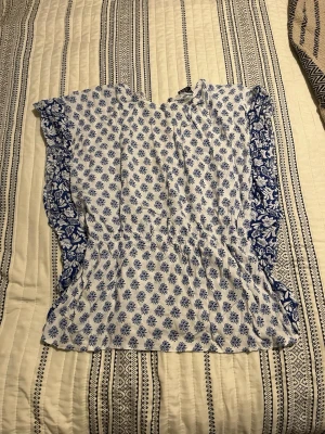 Vit blus med blått blommönster - Söt vit blus från LCW med blått blommönster och volangdetaljer längs sidorna. Blusen har korta ärmar och är tillverkad i ett lätt och luftigt tyg, perfekt för varma dagar. Passar dig som gillar en romantisk och somrig stil.
