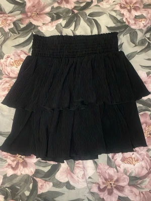 Svart volangkjol från Kappahl - Supersnygg svart kjol från Kappahl med bred smock i midjan och dubbla volanger. Kjolen har en kort modell och är gjord i ett lätt, plisserat tyg som ger en luftig känsla. Perfekt för dig som vill ha en trendig och bekväm look.
