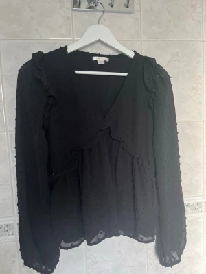 Svart blus med volanger - Snygg svart blus med v-ringning och volangdetaljer. Blusen har långärmad design och är gjord i ett tunt tyg med prickig struktur. Den är i storlek 158/164 men passar XS samt S💓