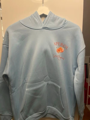 Ljusblå hoodie med Aperol-tryck - Ljusblå hoodie med stor ficka framtill och huva. På bröstet och ryggen finns tryck med Aperol Spritz-motiv och texten 'Aperol' samt illustration av drinkar. Hoodien har en avslappnad passform och är tillverkad i mjukt material, perfekt för en chill stil.