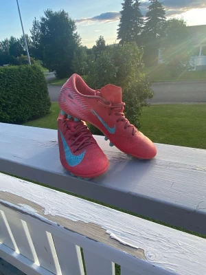 Rosa Nike fotbollsskor med blå detaljer - Säljer ett par snygga fotbollsskor från Nike i en fräsch rosa färg med blå Nike-logga och vita detaljer på sidorna. Skorna har snörning och dobbar för spel på gräs. Perfekta för dig som vill sticka ut på planen!