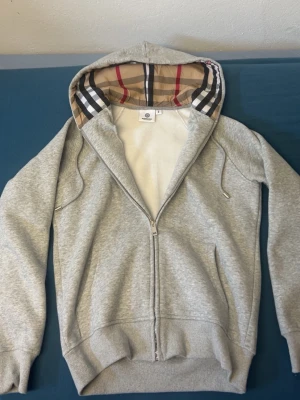 Grå hoodie med Burberry-mönstrad huva - Snygg grå zip-hoodie från Burberry med klassiskt rutigt mönster i huvan. Tröjan har dragsko i huvan, ribbade muddar och två fickor framtill. Perfekt för en chill och stilren look. Materialet är mjukt och bekvämt, troligen bomullsblandning.