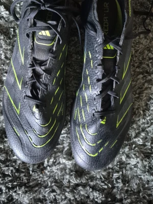 Svarta Adidas Predator fotbollsskor - Säljer ett par svarta Adidas Predator fotbollsskor med neongula detaljer och snörning. Skorna har en modern, strömlinjeformad design och är tillverkade i syntetmaterial för bra grepp och kontroll på planen. Perfekta för dig som vill sticka ut på matchen!