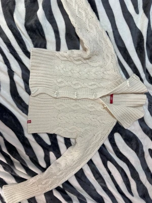 Vit kabelstickad kort kofta från edc - Säljer en mysig vit kabelstickad kofta från edc by esprit. Koftan har stora knappar framtill, ribbade muddar och ett klassiskt flätmönster. Perfekt för lager på lager och ger en chill vibe till din outfit. Den är en kort kofta som passar bra till jeans :) står storlek L men den är liten i storleken så skulle säga S/XS