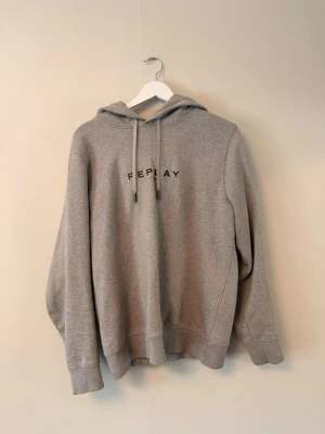 Grå hoodie från Replay - Säljer en stilren grå hoodie från Replay med svart logga på bröstet. Hoodien har huva med dragsko och ribbade muddar vid ärmar och nederkant. Perfekt för en avslappnad och trendig look.  Storleken är mellan S OCH M men är på papper M
